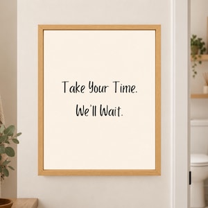 Framed minimalist bathroom wall art with the handwritten black phrase “Take Your Time. We’ll Wait.” centered on a warm off-white background in a light wood frame beside a bathroom doorway.