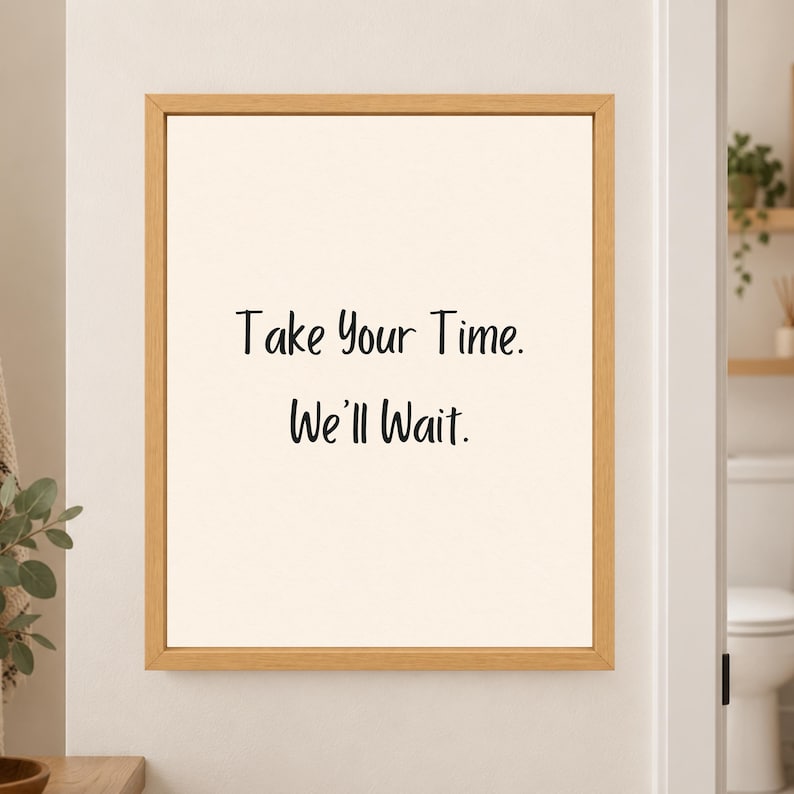 Framed minimalist bathroom wall art with the handwritten black phrase “Take Your Time. We’ll Wait.” centered on a warm off-white background in a light wood frame beside a bathroom doorway.
