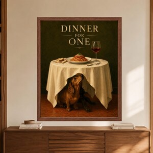 Framed dachshund wall art titled Dinner for One, showing a brown dachshund sitting beneath a white tablecloth with spaghetti, bread and a glass of red wine on the table above.