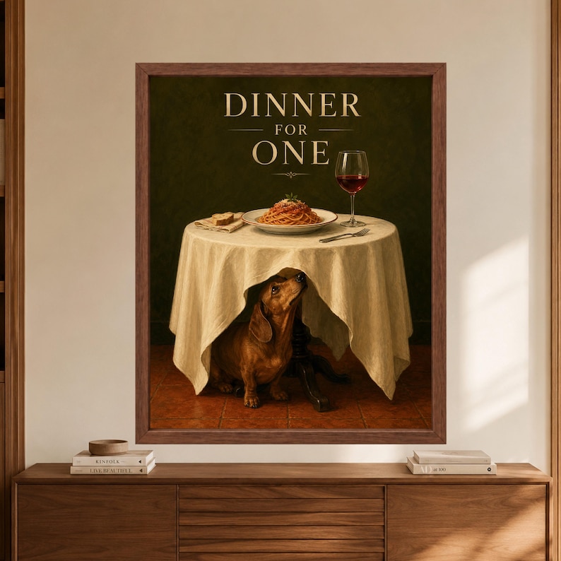 Framed dachshund wall art titled Dinner for One, showing a brown dachshund sitting beneath a white tablecloth with spaghetti, bread and a glass of red wine on the table above.