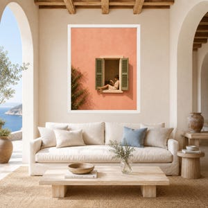 May include: A framed wall art print featuring a peach-colored exterior wall with an open window. Green shutters frame the window, where a person sits reading. The scene is set against a clear blue sky, capturing a Mediterranean architectural style.