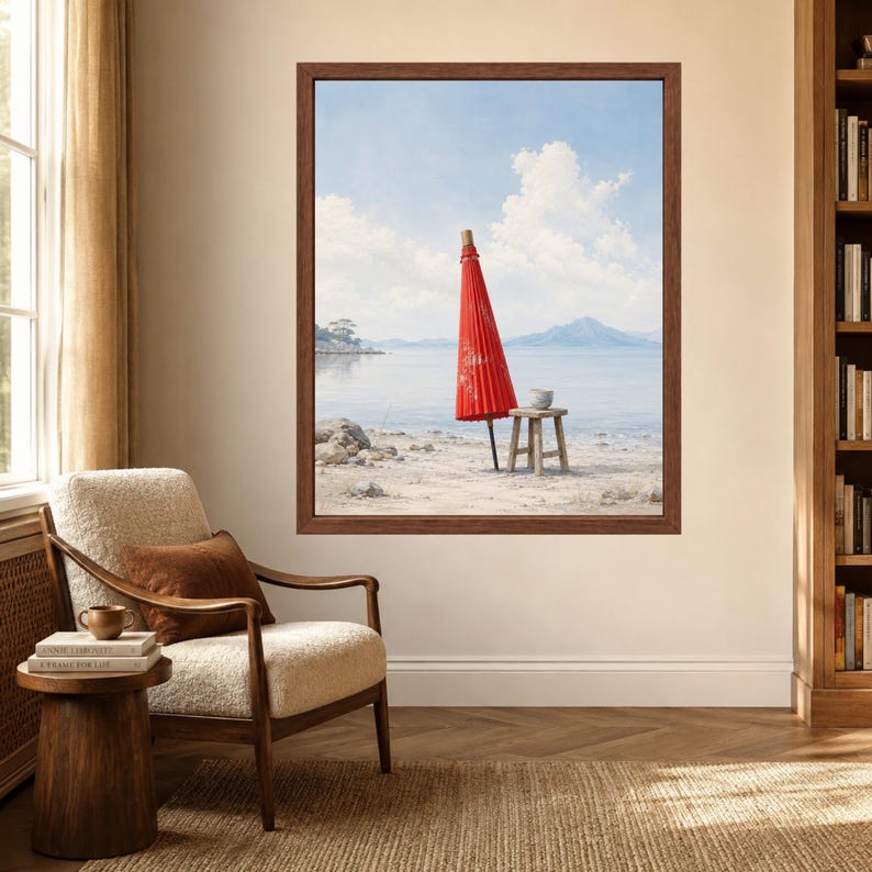 May include: A framed wall art print depicting a bright red paper umbrella standing upright on a sandy beach next to a small wooden stool. The background features a calm body of water, distant mountains, and a light blue sky with white clouds.