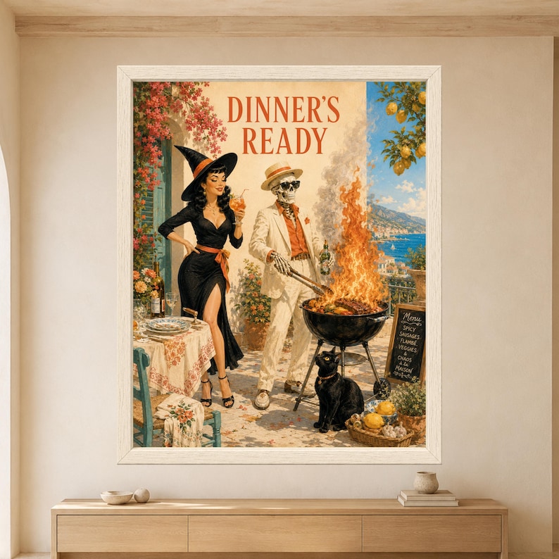 Framed Dinner's Ready wall art showing a skeleton grilling over a huge barbecue flame beside a retro witch holding a cocktail and a black cat on a sunny Mediterranean terrace.
