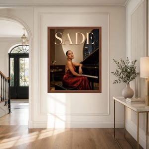 May include: A framed wall art print featuring a person in a dark red satin dress sitting at a black grand piano. The word SADE is printed in large, light-colored capital letters at the top. The scene is set in a dimly lit room with a warm glow.