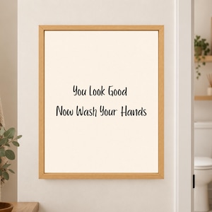 Framed minimalist bathroom wall art with the handwritten black phrase “You Look Good Now Wash Your Hands” centered on a warm off-white background in a light wood frame beside a bathroom doorway.