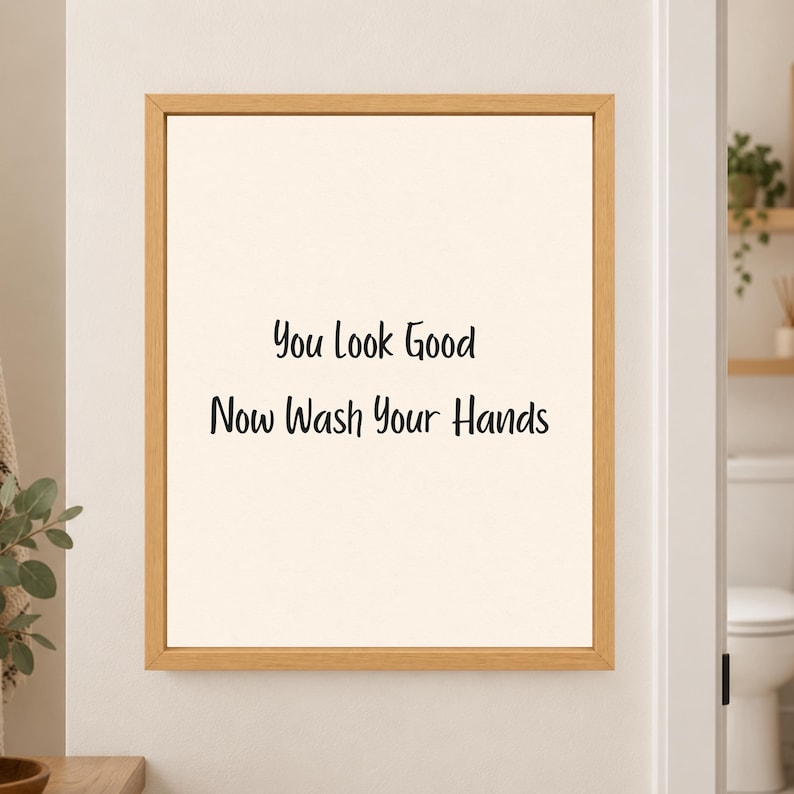 Framed minimalist bathroom wall art with the handwritten black phrase “You Look Good Now Wash Your Hands” centered on a warm off-white background in a light wood frame beside a bathroom doorway.