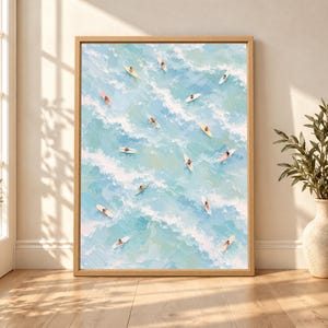 Framed aerial surf wall art showing multiple surfers on pastel surfboards drifting through soft blue ocean waves with white foam in a light textured coastal painting.