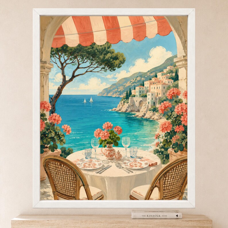 May include: A framed wall art print depicting a coastal scene from a balcony with a red and white striped awning. A round table with two wicker chairs, plates, and glasses overlooks a blue sea, distant sailboats, and a hillside village.