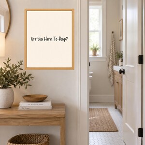 May include: A rectangular wall art piece in a light wood frame featuring a cream-colored background with the black text "Are You Here To Poop?" centered on the print. The artwork is displayed on a wall above a wooden console table.