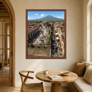 K&ouml;nnte beinhalten: Ein gerahmter Wandkunstdruck, der eine Luftaufnahme der antiken Stadt Pompeji mit dem Vesuv im Hintergrund zeigt. Das Bild zeigt Steingeb&auml;ude, einen zentralen Platz und den in Wei&szlig; am unteren Rand gedruckten Text POMPEII 79 CE.