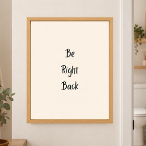 Framed minimalist bathroom wall art with the handwritten black words “Be Right Back” stacked vertically on a warm off-white background in a light wood frame beside a bathroom doorway.