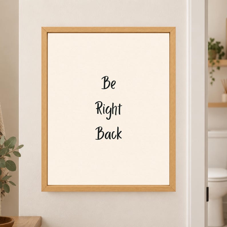 Framed minimalist bathroom wall art with the handwritten black words “Be Right Back” stacked vertically on a warm off-white background in a light wood frame beside a bathroom doorway.