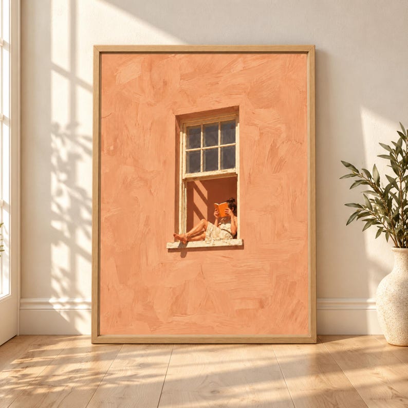 Framed peach wall art showing a woman sitting in an open window reading an orange book against a textured terracotta facade with warm sunlight and minimalist composition.