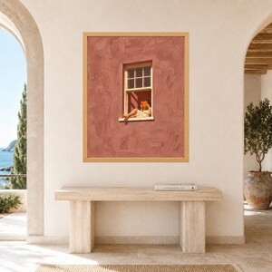 May include: A framed wall art print featuring a textured terracotta-colored wall with a single window. A person sits in the window frame reading an orange book, wearing a light-colored dress. The artwork is enclosed in a light wood frame.