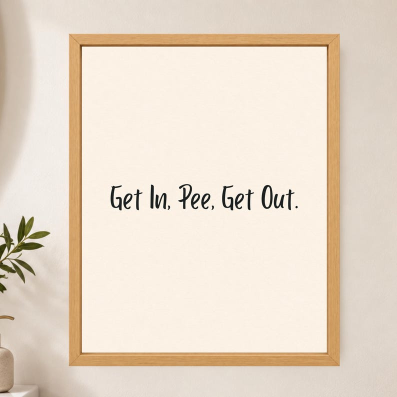 May include: A framed wall art piece featuring a light cream background with the black text Get In, Pee, Get Out. centered in the middle. The artwork is enclosed in a simple, light-toned wooden frame with a clean, rectangular border design.