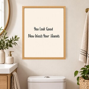 May include: A rectangular wall art piece featuring a light beige background and a thin wooden frame. The text "You Look Good Now Wash Your Hands" is printed in black, sans-serif lettering in the center of the print, creating a clear visual statement.
