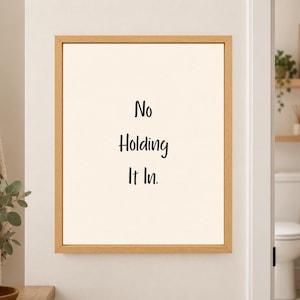 Framed minimalist bathroom wall art with the handwritten black phrase “No Holding It In.” stacked vertically on a warm off-white background in a light wood frame beside a bathroom doorway.