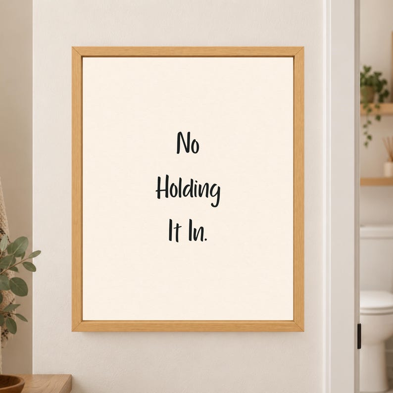 Framed minimalist bathroom wall art with the handwritten black phrase “No Holding It In.” stacked vertically on a warm off-white background in a light wood frame beside a bathroom doorway.