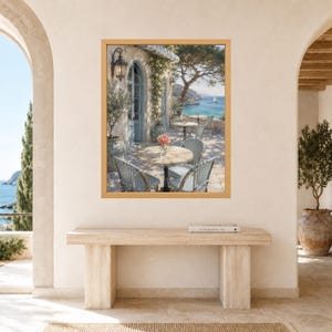 May include: A framed wall art print depicting a Mediterranean seaside terrace with a round stone table, woven chairs, and a vase of pink flowers. The scene features a stone building with an arched doorway, climbing greenery, and a view of the blue sea.