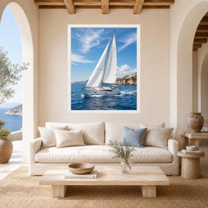 Puede incluir: Una impresi&oacute;n art&iacute;stica enmarcada que muestra un velero blanco con las velas desplegadas navegando en aguas oce&aacute;nicas de color azul profundo bajo un cielo brillante con nubes tenues, exhibida sobre un sof&aacute; beige con cojines neutros.