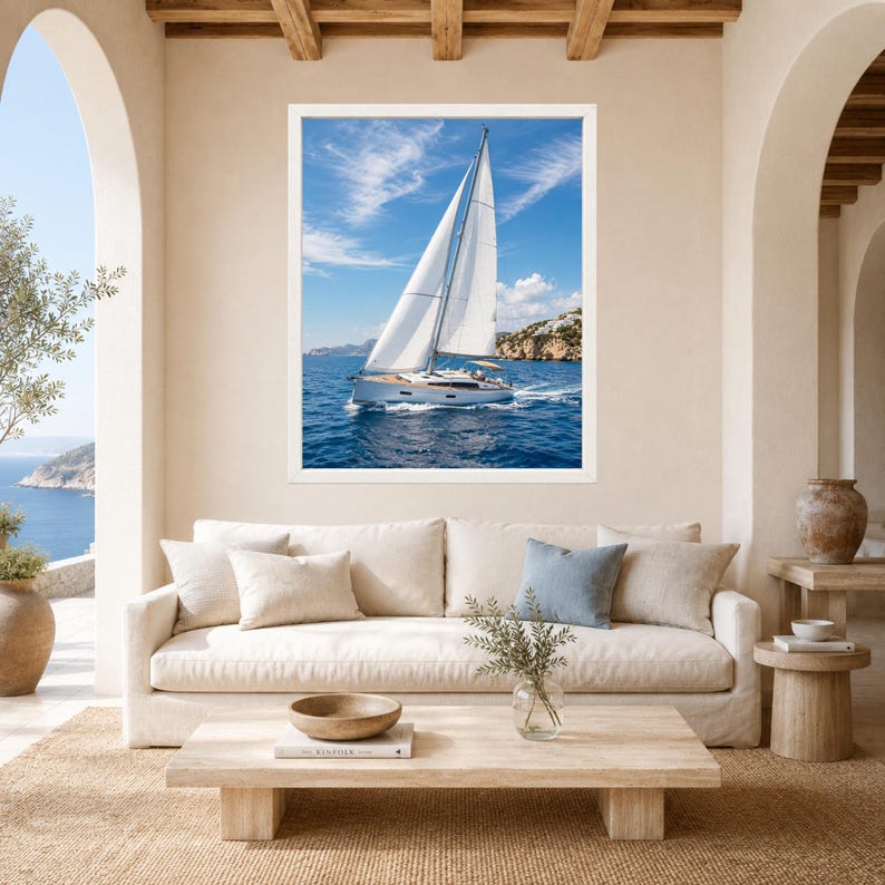 Puede incluir: Una impresi&oacute;n art&iacute;stica enmarcada que muestra un velero blanco con las velas desplegadas navegando en aguas oce&aacute;nicas de color azul profundo bajo un cielo brillante con nubes tenues, exhibida sobre un sof&aacute; beige con cojines neutros.