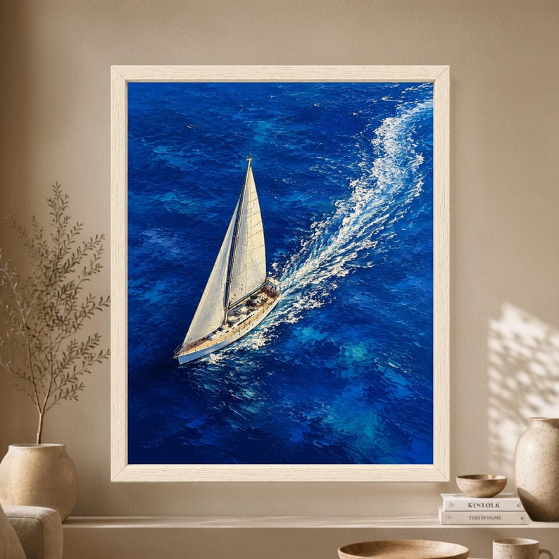 Framed aerial sailboat wall art showing a white sailing yacht crossing deep cobalt blue ocean water from above, with a long curved trail of white foam behind the boat.