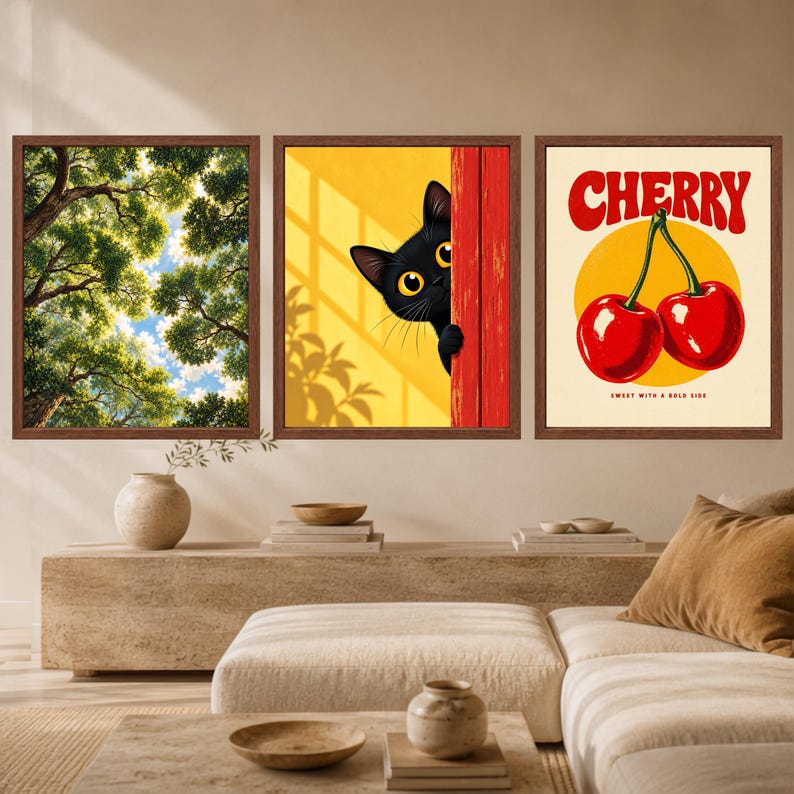 May include: Three framed wall art prints. The left print shows a low-angle view of green tree branches against a blue sky. The centre print features a black cat peeking from behind a red door against a yellow background. The right print displays two red cherries with the text CHERRY and SWEET WITH A BOLD SIDE.
