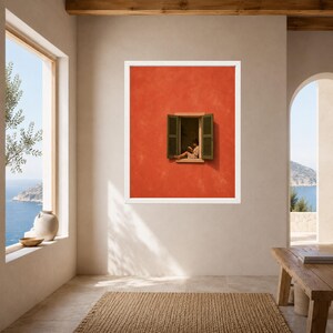 May include: A framed wall art print featuring a vibrant orange wall with a centred window. The window has open dark green shutters and a person sitting on the ledge reading a book. The artwork is displayed in a white frame on a neutral wall.