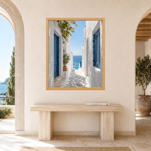 K&ouml;nnte beinhalten: Ein gerahmter Kunstdruck, der eine schmale wei&szlig;e Gasse mit leuchtend blauen T&uuml;ren und Fensterl&auml;den zeigt, die den Blick auf das blaue Meer unter klarem Himmel freigibt. Die Szene umfasst wei&szlig;en Steinpflasterboden, eine Topfpflanze und wei&szlig;e Ranken.