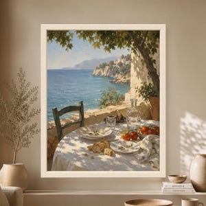 Framed Mediterranean dining wall art showing a rustic seaside table with tomatoes, bread, ceramic plates and a glass carafe overlooking a blue coastal bay and distant hillside village.