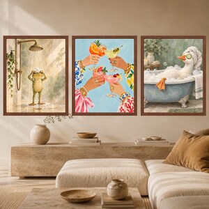 May include: A set of three framed wall art prints. The left print shows a frog showering. The center print depicts four hands holding colorful cocktails with fruit garnishes. The right print features a white duck in a bubble bath with cucumber slices.