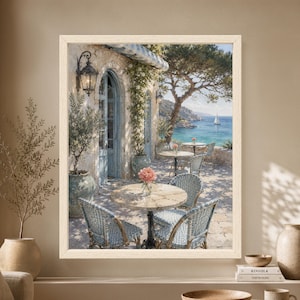 Framed Mediterranean cafe wall art showing a sunlit stone seaside terrace with blue woven bistro chairs, olive trees, flowering vines and a sailboat crossing the clear blue coastal water.