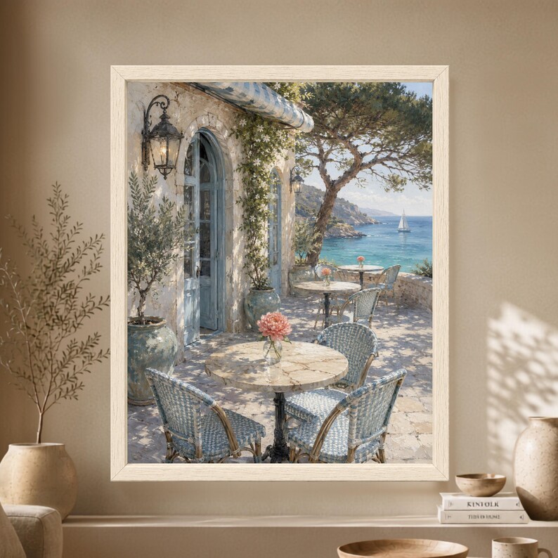 Framed Mediterranean cafe wall art showing a sunlit stone seaside terrace with blue woven bistro chairs, olive trees, flowering vines and a sailboat crossing the clear blue coastal water.