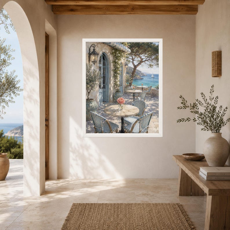 May include: A framed wall art print depicting a Mediterranean seaside terrace with round tables, woven chairs, and a vase of pink flowers. The scene features a stone building with a blue arched doorway, a lantern, and a view of the blue sea.