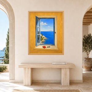May include: A framed wall art print depicting an open blue window frame set into a textured yellow wall. The view through the window shows a bright blue sea, a rocky coastline, and a clear sky. A small red book and black sunglasses sit on the sill.