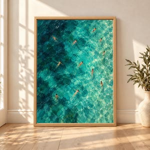 Framed aerial ocean wall art showing multiple swimmers floating across clear turquoise, aqua and teal water in a textured painterly coastal composition.