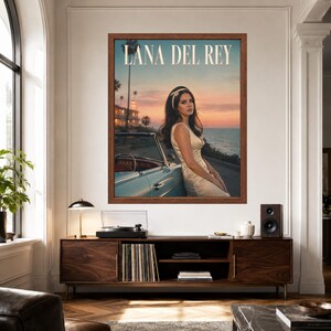 May include: A framed wall poster featuring Lana Del Rey leaning against a light blue vintage convertible car at sunset by the ocean. The text LANA DEL REY is printed in large white capital letters at the top of the image, which shows a coastal road.