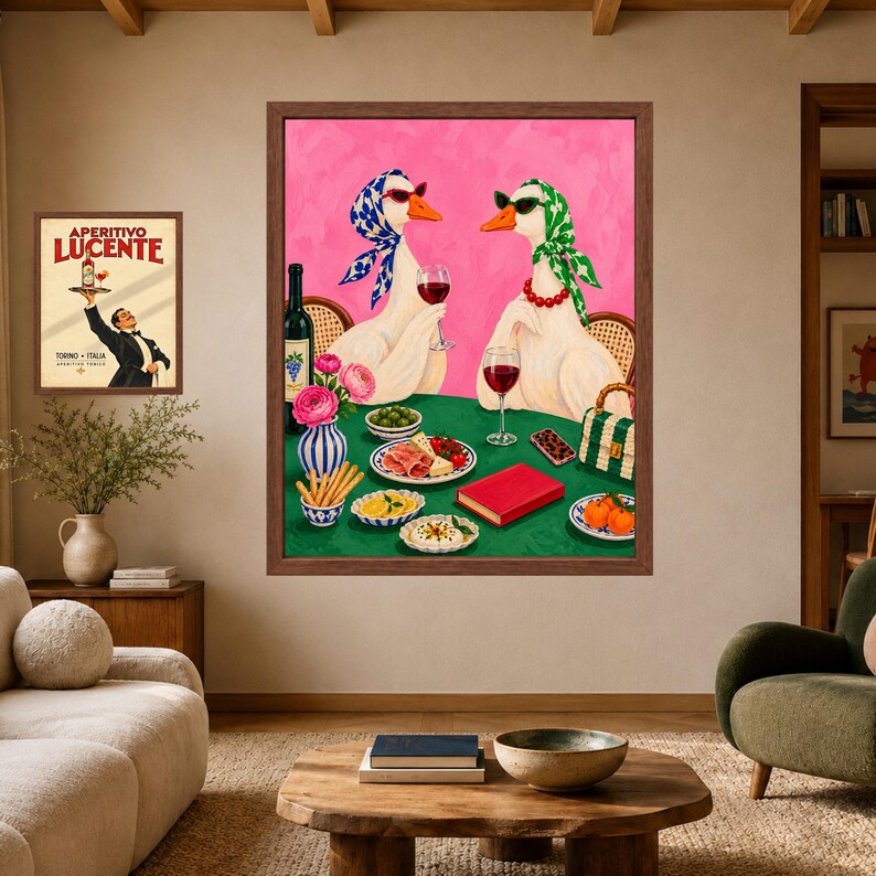May include: A framed wall art print featuring two white geese wearing headscarves and sunglasses, sitting at a green table with wine, snacks, and a red book. A smaller framed poster shows a man holding a bottle labelled Aperitivo Lucente.