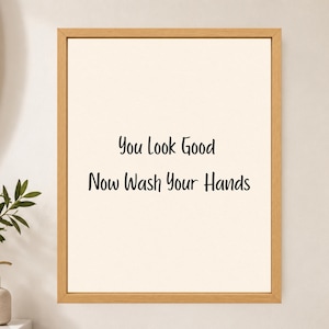 May include: A rectangular wall art piece featuring a light cream background enclosed in a natural light-toned wooden frame. The center displays black text that reads: You Look Good Now Wash Your Hands, written in a simple, clean, sans-serif font.