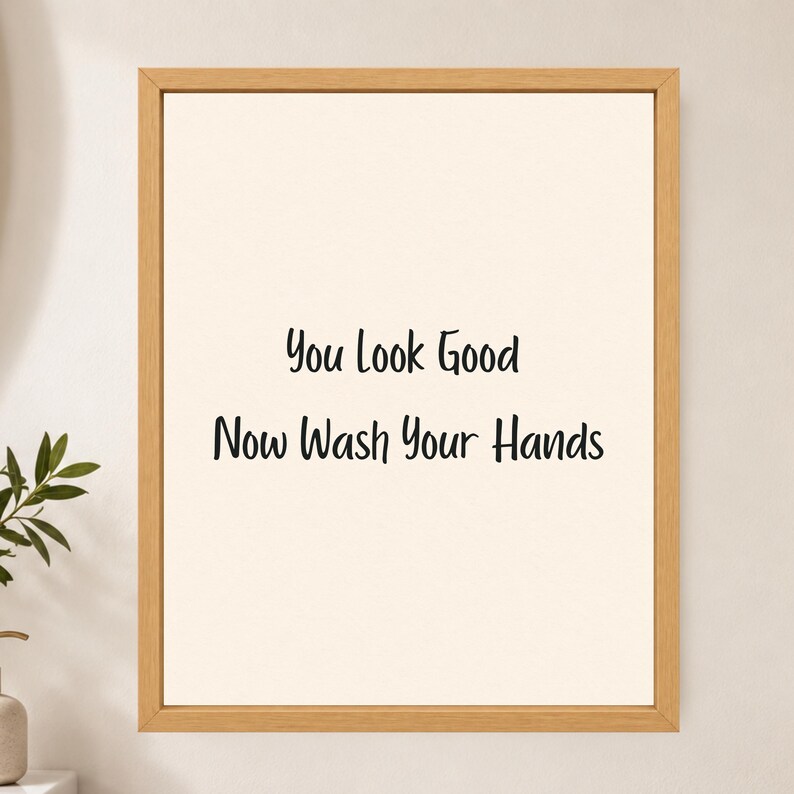 May include: A rectangular wall art piece featuring a light cream background enclosed in a natural light-toned wooden frame. The center displays black text that reads: You Look Good Now Wash Your Hands, written in a simple, clean, sans-serif font.