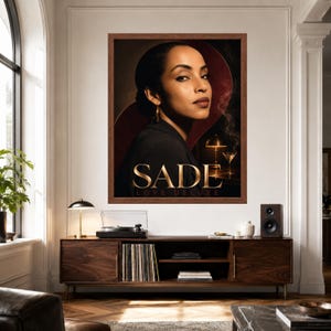 K&ouml;nnte beinhalten: Ein gerahmtes Wandposter mit einem Portr&auml;t von Sade und dem Text SADE LOVE DELUXE. Das Kunstwerk zeigt eine Person in einem dunklen Blazer vor einem dunklen Hintergrund mit einer leuchtenden Lampe und einem Martiniglas. Das Poster h&auml;ngt an einer wei&szlig;en Wand.