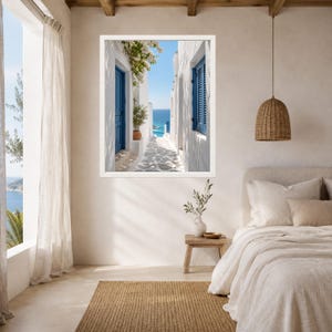 K&ouml;nnte beinhalten: Ein gerahmter Kunstdruck, der eine schmale wei&szlig;e Gasse in einer mediterranen K&uuml;stenstadt zeigt, mit leuchtend blauen T&uuml;ren und Fensterl&auml;den, einer Topfpflanze und einem Blick auf das blaue Meer unter klarem Himmel, in einem Schlafzimmer.