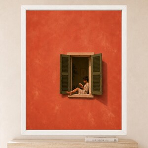 May include: A framed wall art print featuring a vibrant orange textured wall with an open window. Inside the window frame, a figure sits on the ledge reading a book. The window has dark green louvered shutters on both sides, set against the wall.