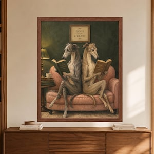 Framed greyhound reading wall art showing two sighthounds seated on a dusty pink sofa with open books beneath a Dogs at the Library sign, beside a lamp, coffee cup and stacked books on a dark green background.