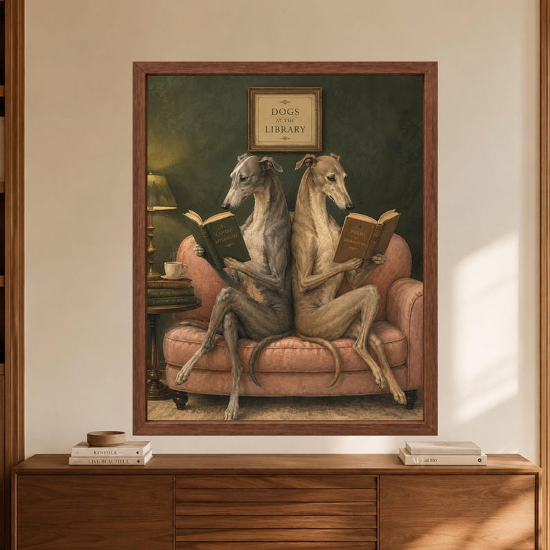 Framed greyhound reading wall art showing two sighthounds seated on a dusty pink sofa with open books beneath a Dogs at the Library sign, beside a lamp, coffee cup and stacked books on a dark green background.