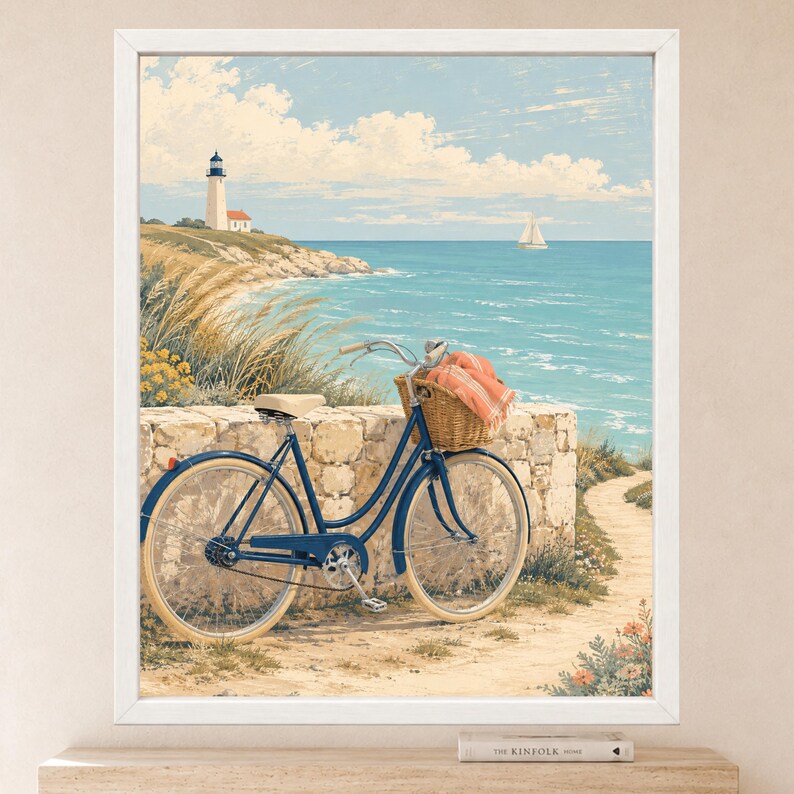 Op de afbeelding: Een ingelijste kunstdruk met een kusttafereel en een blauwe fiets tegen een stenen muur. De fiets heeft een rieten mand met een koraalkleurige doek. Op de achtergrond staat een vuurtoren op een klif aan de zee.