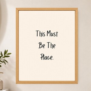 May include: A framed wall art piece featuring a light cream background with the black text "This Must Be The Place." centered in the frame. The artwork is enclosed in a simple, light-toned wooden frame with a clean, rectangular profile.