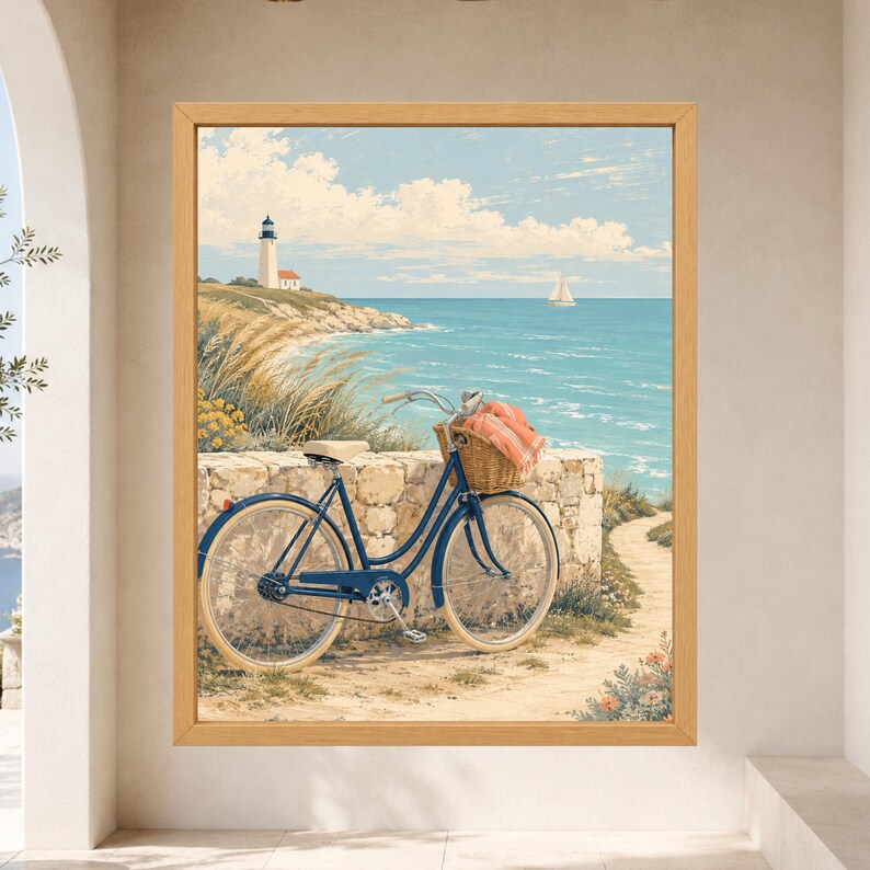Op de afbeelding: Een ingelijste kunstdruk met een kusttafereel met een blauwe fiets geparkeerd tegen een stenen muur. De fiets heeft een rieten mand met een gevouwen oranje doek. Op de achtergrond staat een vuurtoren op een grasachtige klif die uitkijkt over de blauwe oceaan met een zeilboot zichtbaar aan de horizon onder een heldere lucht.