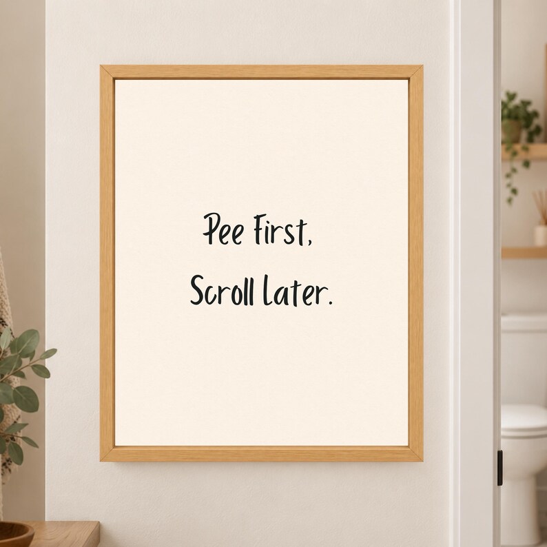 Framed minimalist bathroom wall art with the handwritten black phrase “Pee First. Scroll Later.” centered on a warm off-white background in a light wood frame beside a bathroom doorway.
