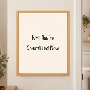 Framed minimalist bathroom wall art with the handwritten black phrase “Well, You’re Committed Now.” centered on a warm off-white background in a light wood frame beside a bathroom doorway.